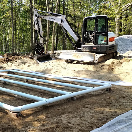 A septic system installation service is in progress in the dirt area cleared by an excavator.